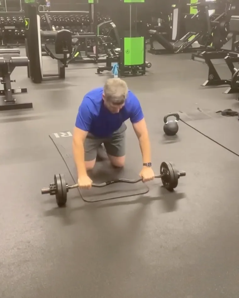 Charlie kneeling on a mat, gripping an EZ bar at the start of a clean, a kettlebell beside him