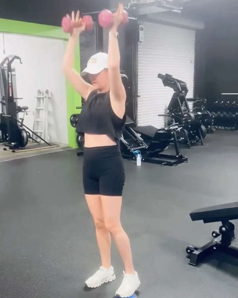 Sara pressing two dumbbells overhead, one foot resting on a slider pad
