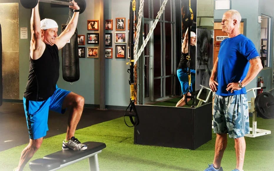 An older client stepping up onto a bench while pressing a barbell overhead, with personal trainer Craig Chapin watching the rep