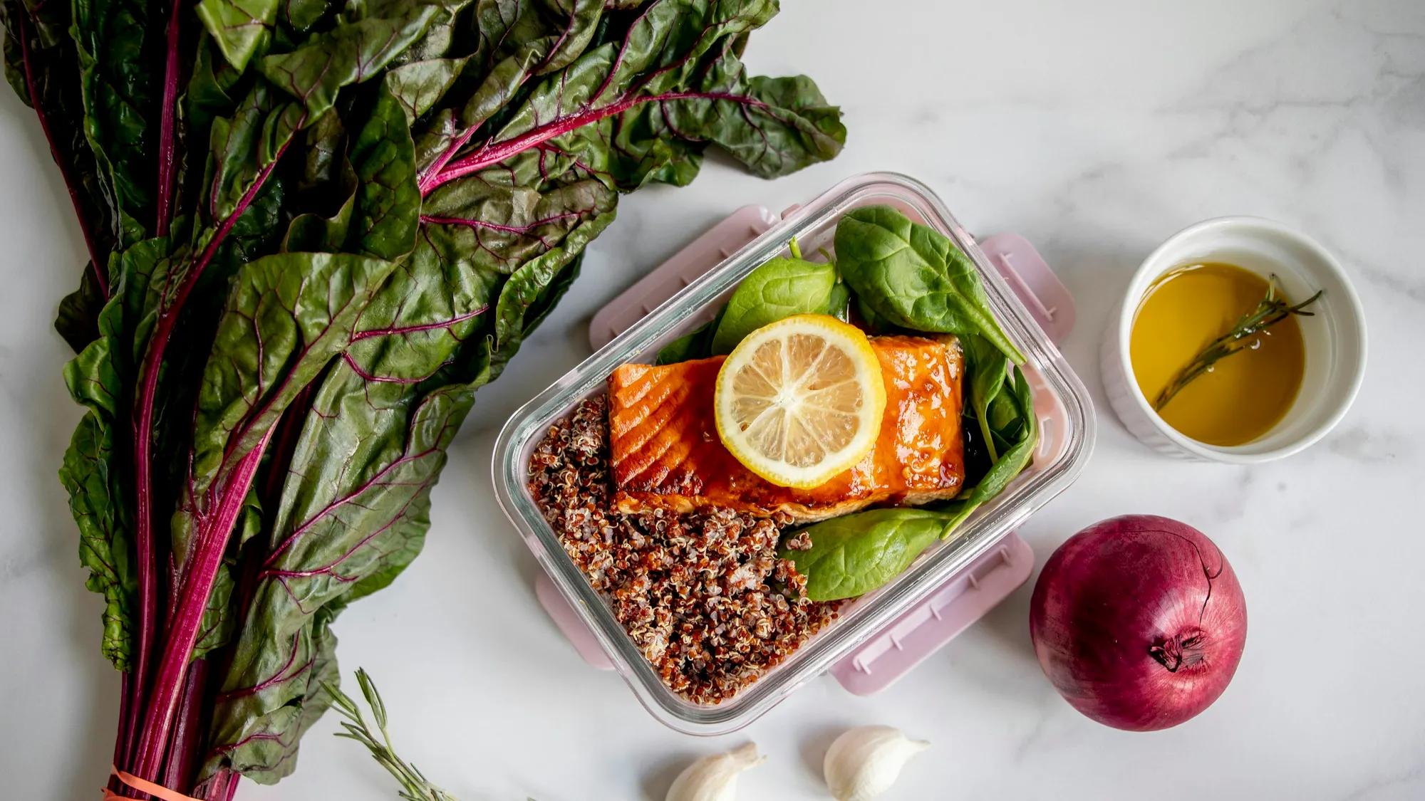 A glass meal-prep box of salmon, quinoa and spinach on a marble counter beside rainbow chard, garlic and a red onion