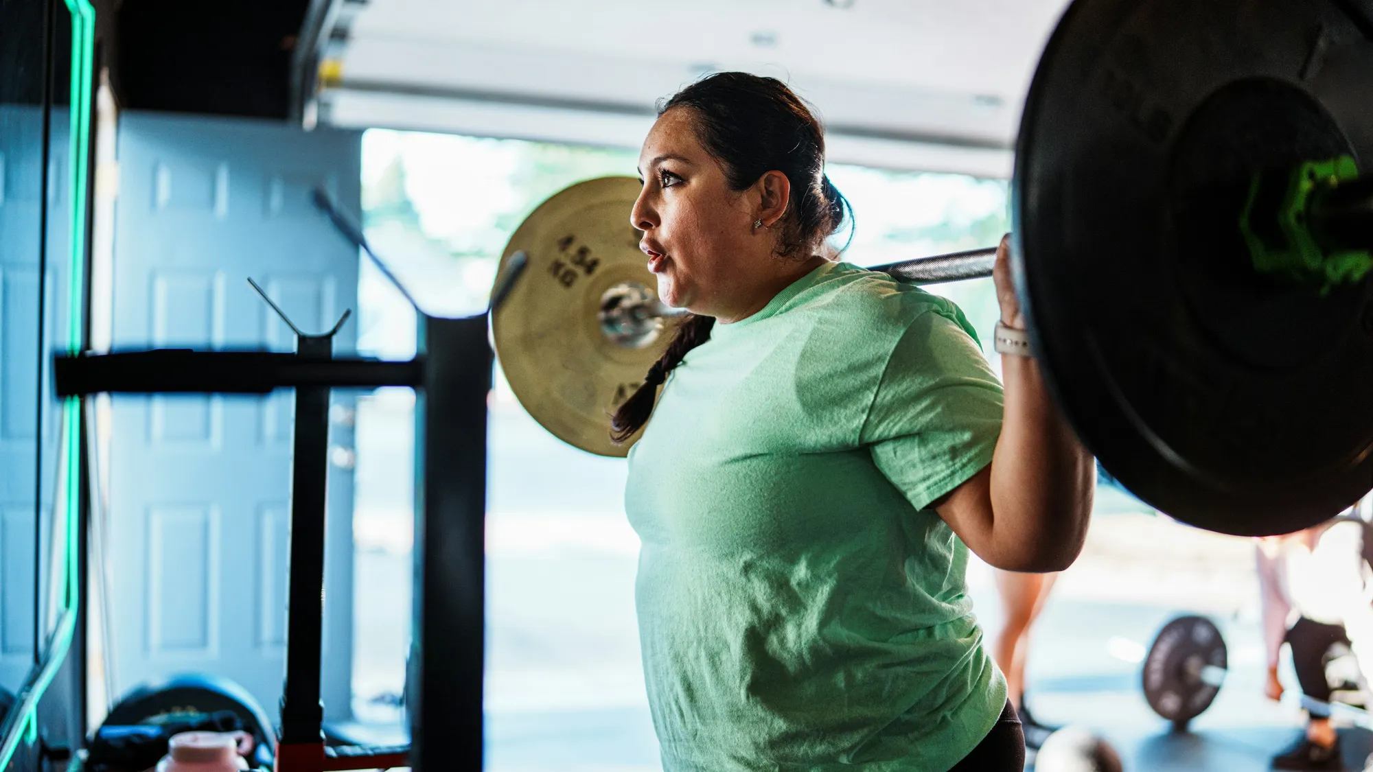 A woman in a green T-shirt braced under a loaded barbell for a back squat, strength training at a weight that is heavy for her