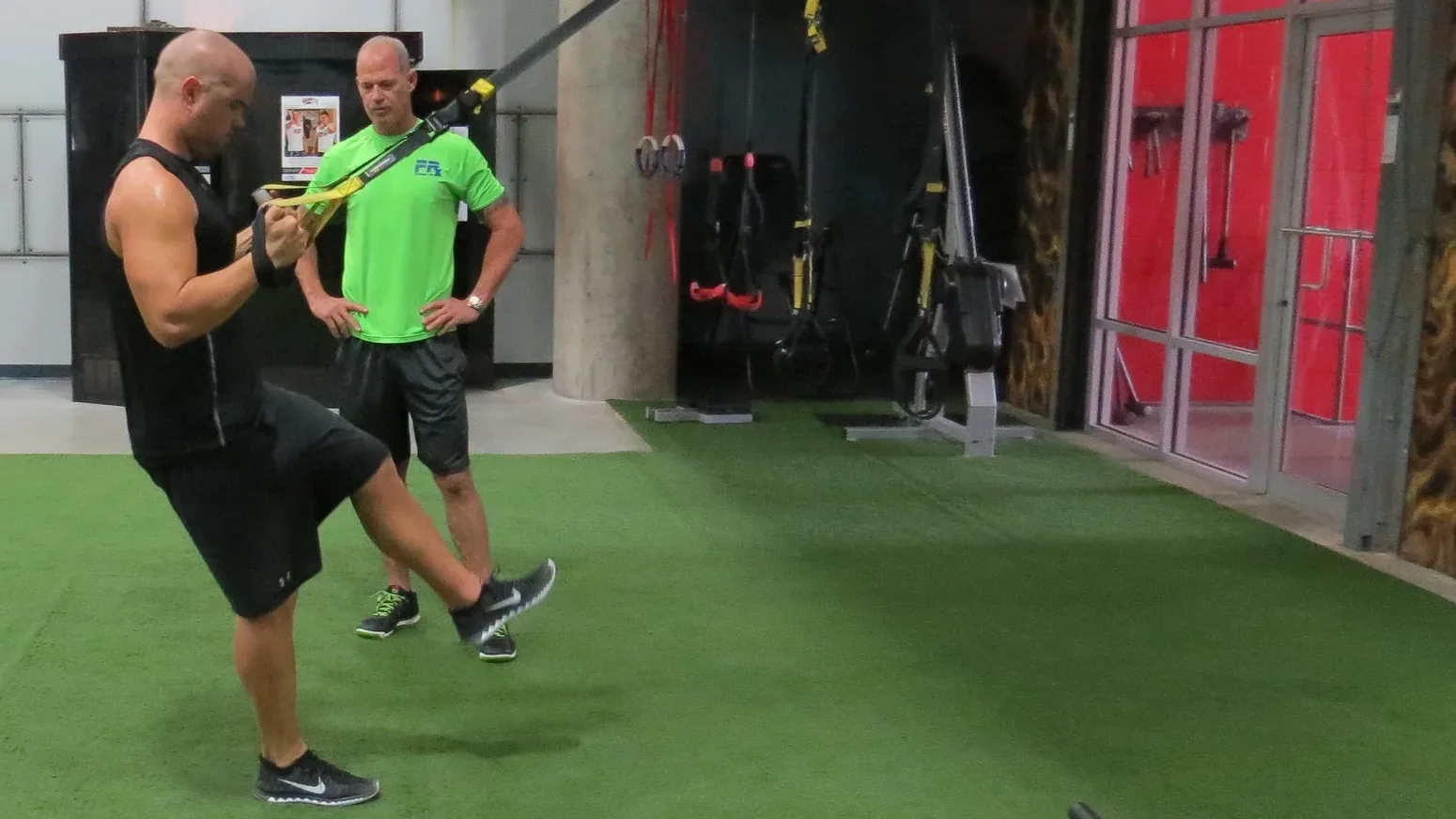 Craig Chapin, a personal trainer for men over 50 in South Tampa, watching a male client's form through a single-leg TRX exercise on the training turf