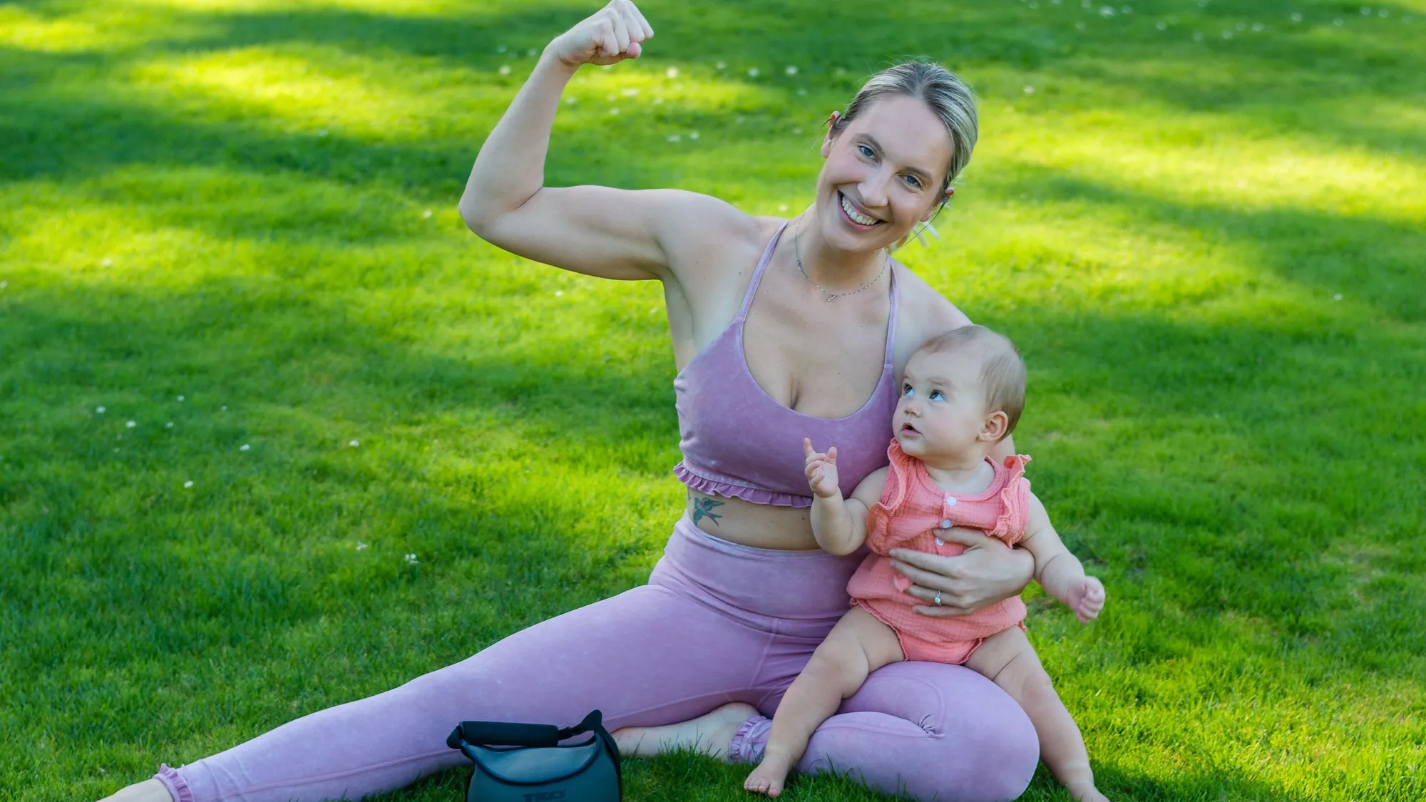 A smiling mother sitting on a sunny lawn with her baby on her lap, one arm raised in a flex
