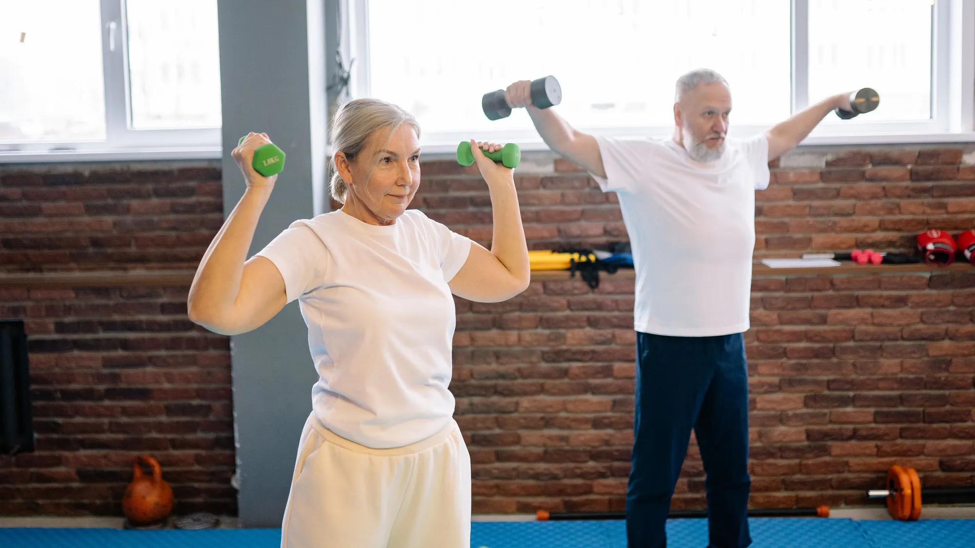 An older woman and man pressing light dumbbells overhead in a bright gym, the kind of strength and balance work senior personal training starts with