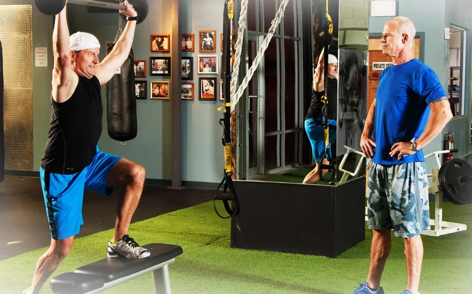 Craig Chapin watching a client press a barbell overhead in a step-up, on the gym floor beside the heavy bags