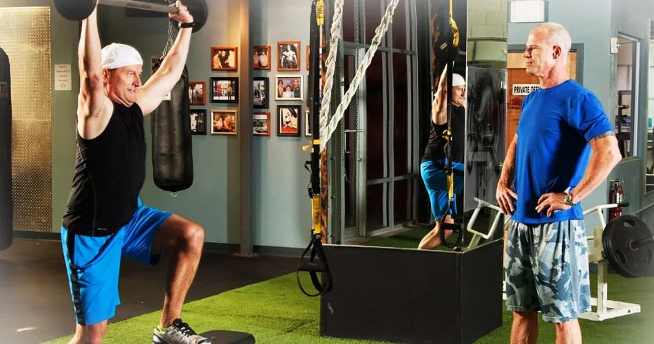 Craig Chapin watching a client hold a barbell locked out overhead at the top of a step-up
