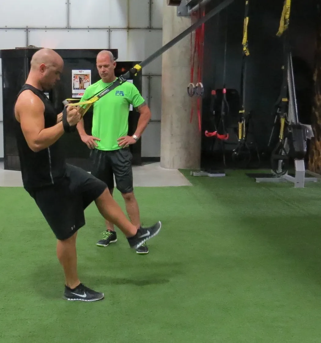 A man mid-rep on a TRX strap, one knee raised, with Craig Chapin standing alongside in a Fitness RX shirt watching his form