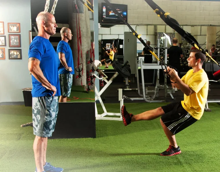 Craig Chapin standing by with his hands behind his back, watching a client lean back into a TRX row