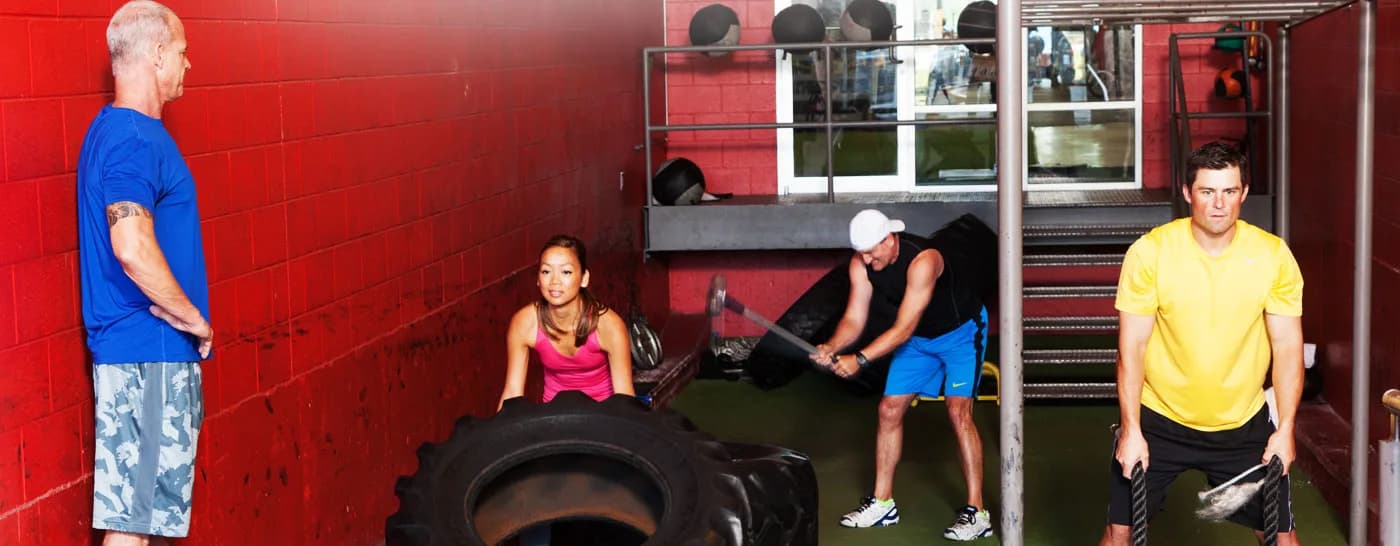 Three clients working in a small group, one flipping a heavy tyre and two on sledgehammers, with Craig Chapin watching from the wall