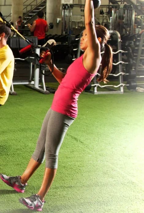 A woman leaning back into a TRX strap mid-row, arms extended overhead, on the training turf