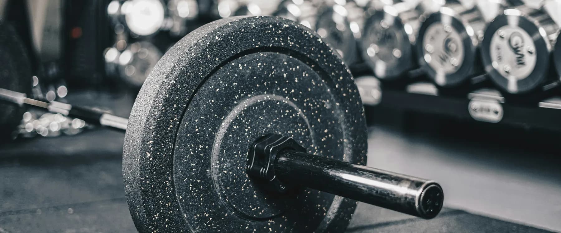 A loaded barbell on the floor of a gym, one bumper plate close to the camera and the bar running out of frame past a rack of dumbbells