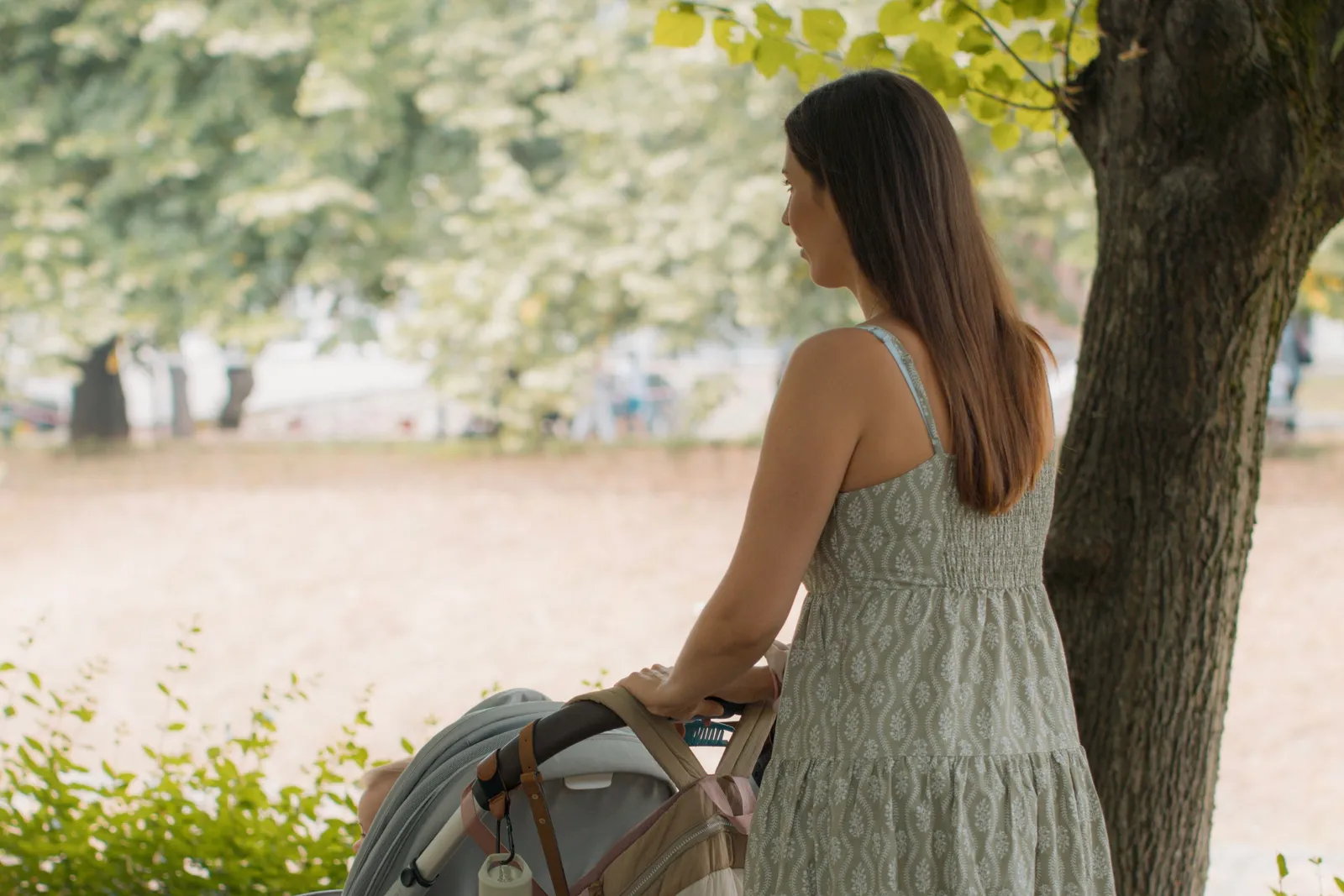 A woman walking a pram along a shaded path through a summer park, her hand resting on the handle