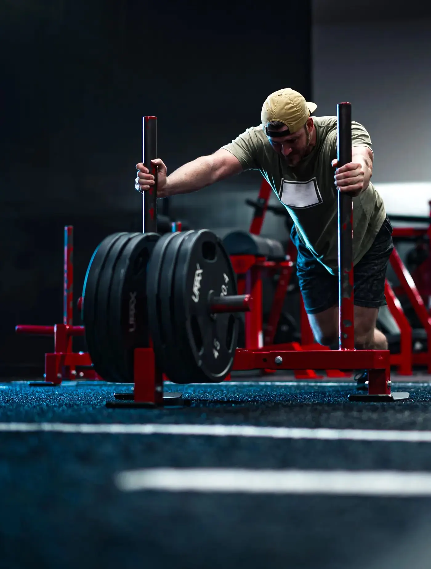 A man driving a weighted sled down a turf lane in a gym, head down over the handles mid-push
